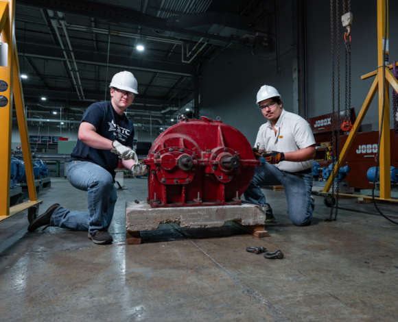 two advanced manufacturing students wearing white helmets and gloves working on a machine
