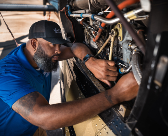 A bearded man leans into the exposed machinery beneath an airplane, possibly making repairs