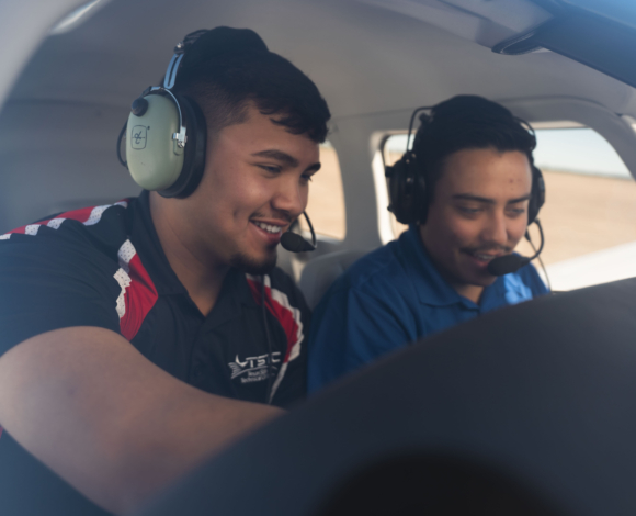 Two people sit in the cockpit of a small airplane. They're wearing headsets and leaning in towards a console, smiling.