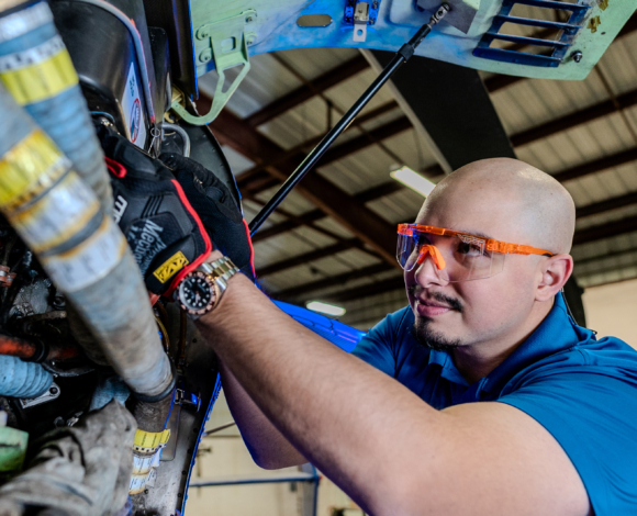 A man in safety glasses reaches into the exposed machinery of an aircraft.