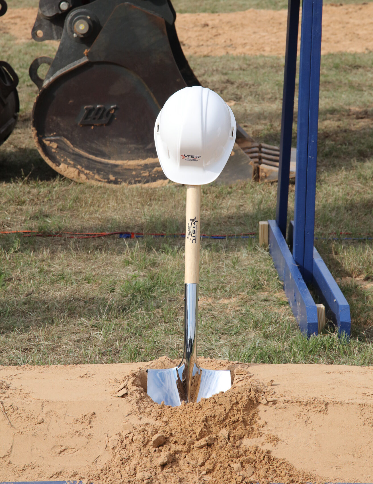A TSTC branded shovel sits in dirt with a white hard hat resting on top of it.